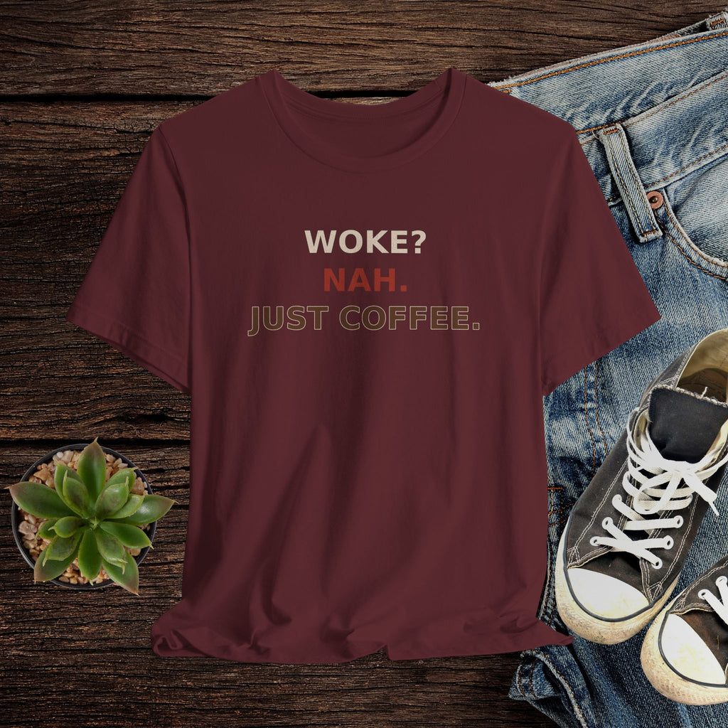 Maroon t-shirt with text 'WOKE? NAH. JUST COFFEE.' on a wooden surface with jeans and sneakers.