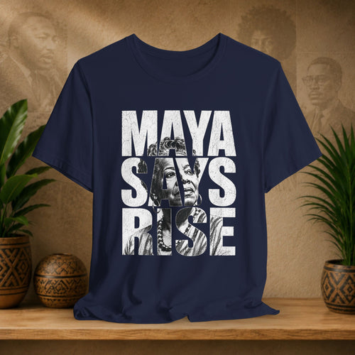 Navy blue t-shirt with 'MAYA SAYS RISE' text and graphic on a wooden surface with plants and decorative items.