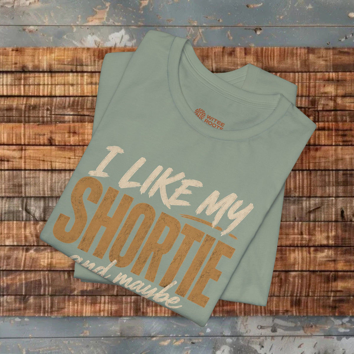 A folded sage green t-shirt with a graphic print reading 'I LIKE MY SHORTIE and maybe' in white and brown lettering, displayed on a rustic wooden plank background. The brand 'White Fox' is visible on the inner neck label.