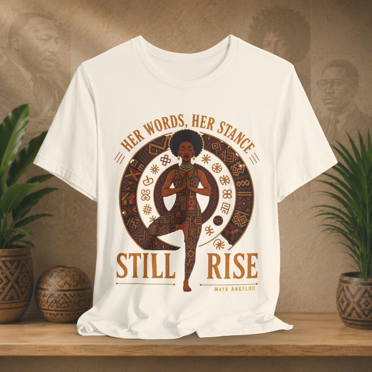 A beige short-sleeve graphic T-shirt displayed on a wooden surface with potted plants in the background. The shirt features a large circular design with a stylized figure in a yoga pose, surrounded by the text 'HER WORDS, HER STANCE STILL RISE' and 'MAYA ANGELOU'.
