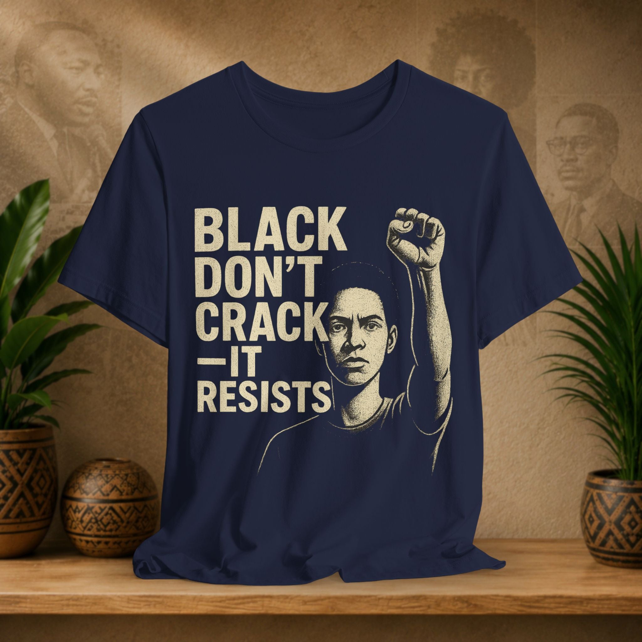 Navy blue t-shirt with graphic and text 'Black Don't Crack, It Resists' on a wooden surface with plants and decorative items.