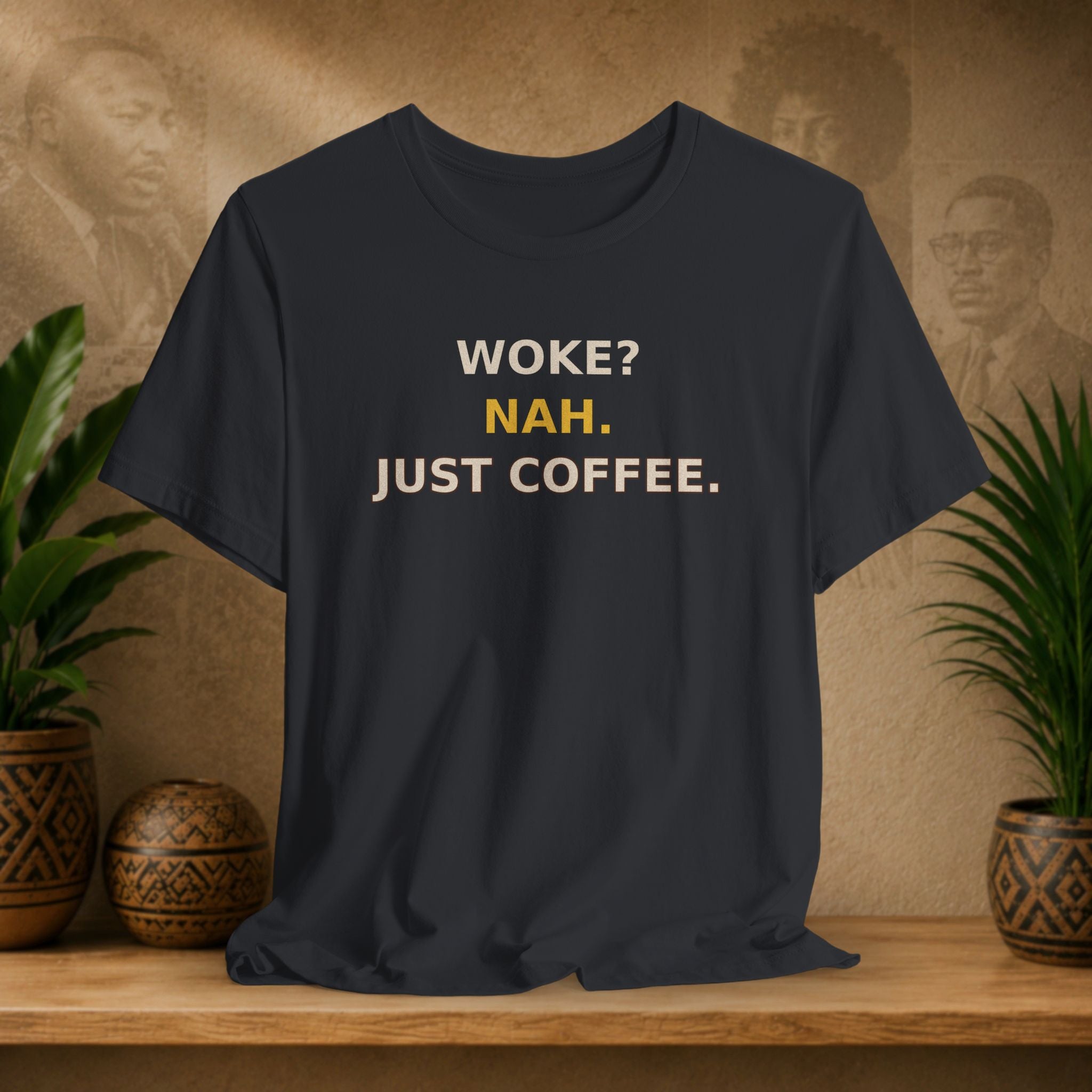Black t-shirt with 'WOKE? NAH. JUST COFFEE.' text on a wooden surface with plants and a textured wall background.