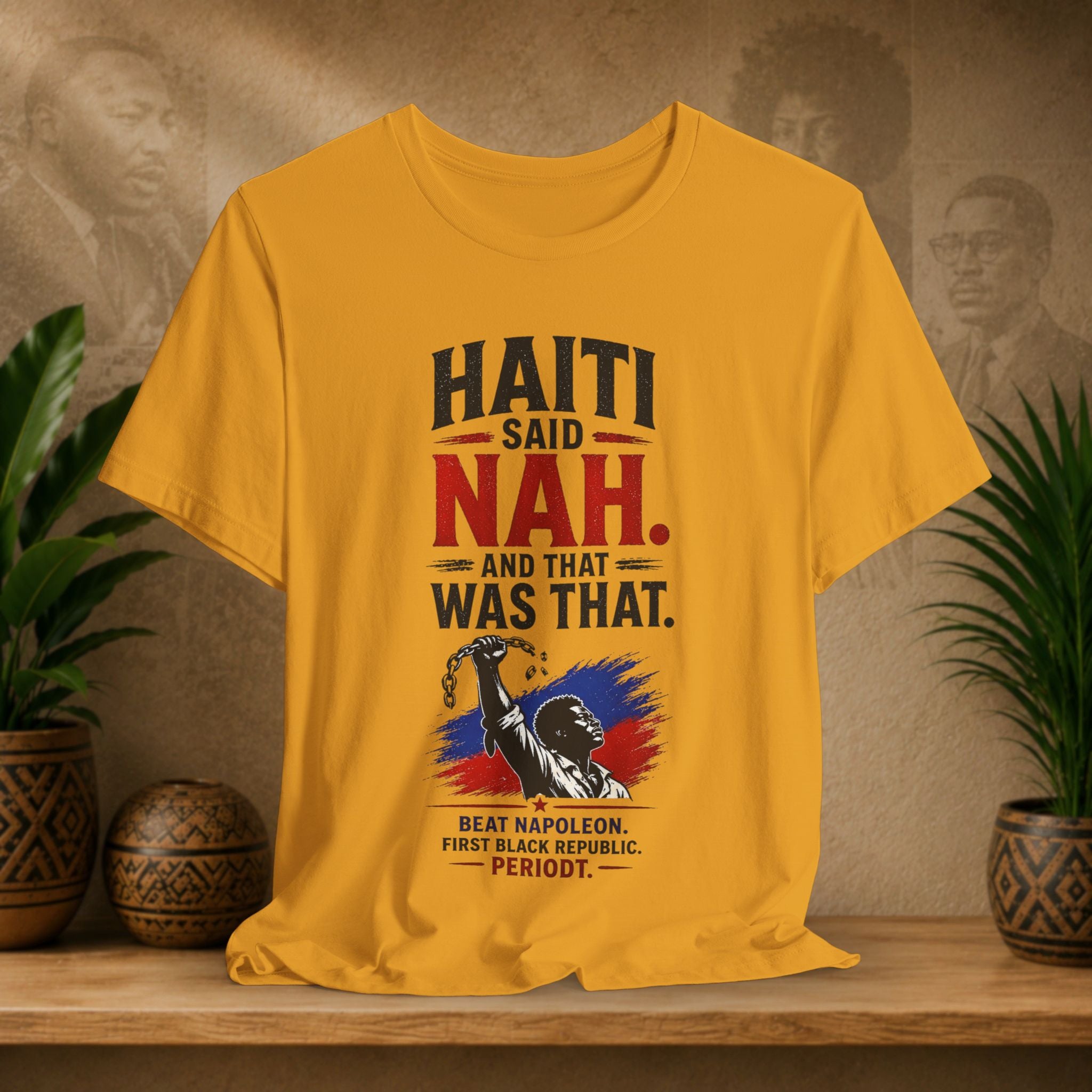 Yellow t-shirt with Haitian text and graphics on a wooden surface with plants and a textured wall background.