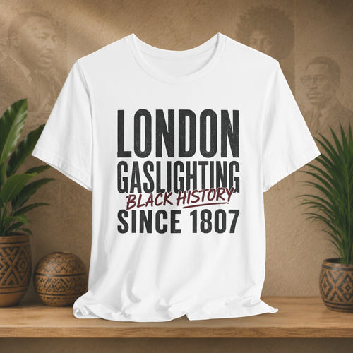 White t-shirt with 'London Gaslighting Black History Since 1807' text on a wooden surface with plants and a blurred background.