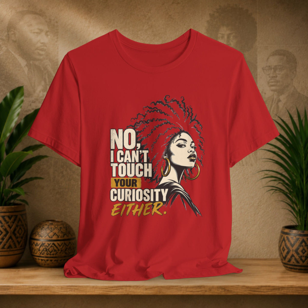 A red short-sleeve graphic T-shirt displayed on a wooden surface with potted plants and a textured wall behind it. The shirt features a stylized illustration of a woman with an afro and bold text that reads, 'NO, I CAN'T TOUCH YOUR CURIOSITY EITHER.
