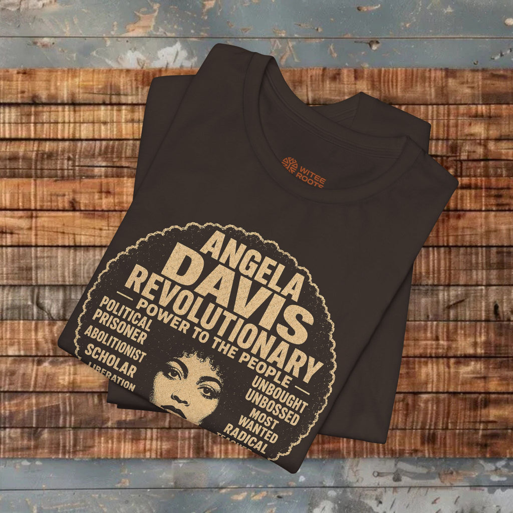A folded brown t-shirt with a vintage-style graphic of Angela Davis, displayed on a rustic wooden plank surface. The graphic features beige text and an illustration of Angela Davis's face, with the words 'ANGELA DAVIS REVOLUTIONARY' and 'POWER TO THE PEOPLE'.