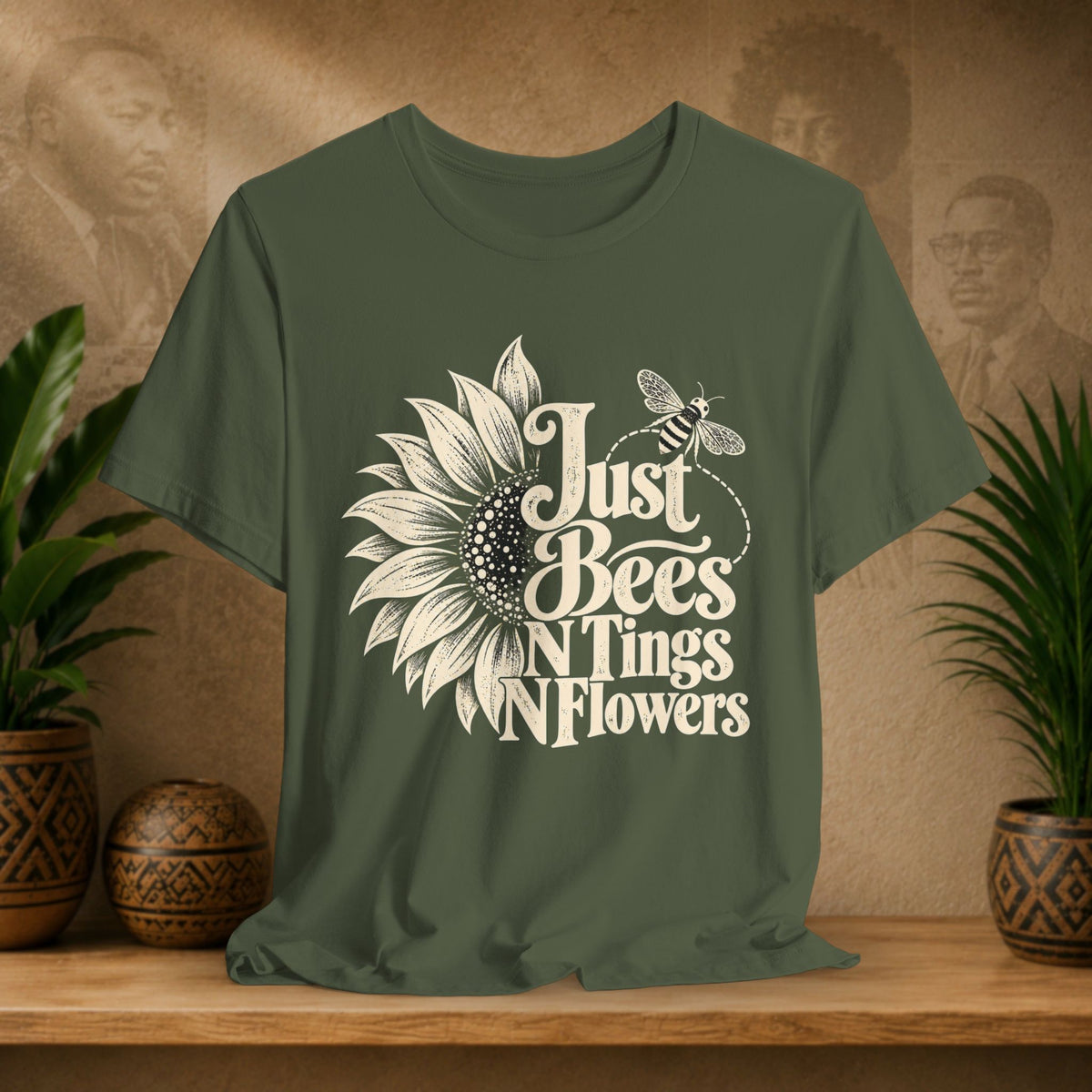 An olive green short-sleeve t-shirt with a white graphic print reading 'Just Bees N' Flowers' alongside a sunflower and bee illustration, displayed on a wooden shelf with potted plants and a textured background.