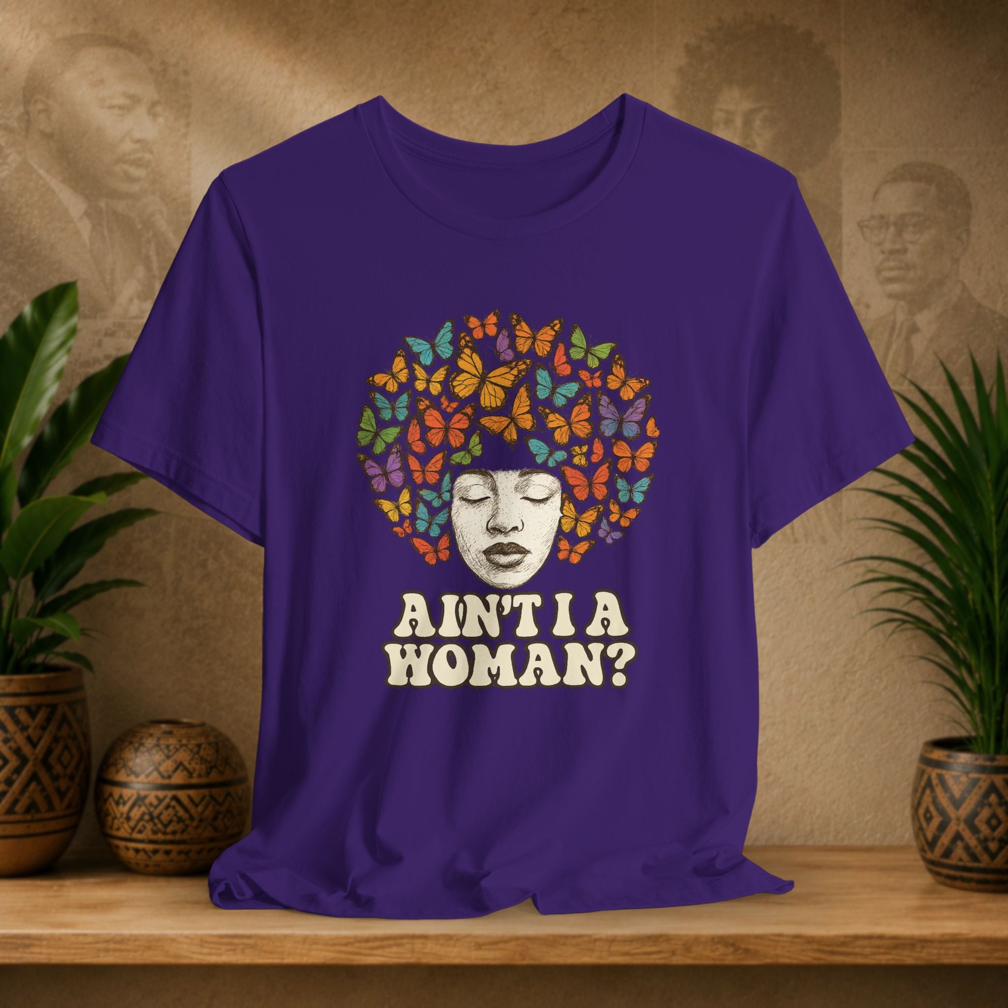 A purple short-sleeve graphic t-shirt displayed on a wooden shelf. The shirt features a graphic of a Black woman's face surrounded by a vibrant multicolor afro made of butterflies, with the text 'AIN'T I A WOMAN?' printed below in white.