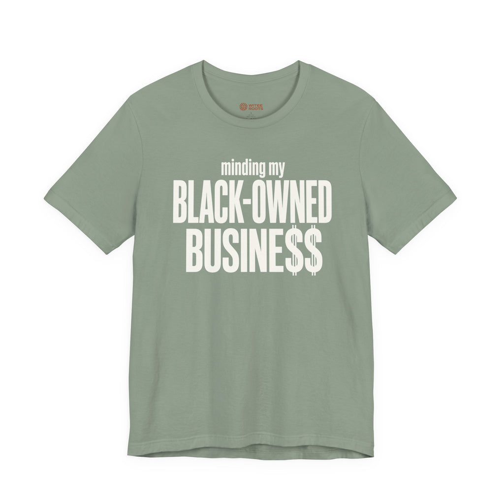A sage green crewneck t-shirt with a white graphic text print reading 'minding my BLACK-OWNED BUSINESS$$' on the front, displayed against a solid white background.