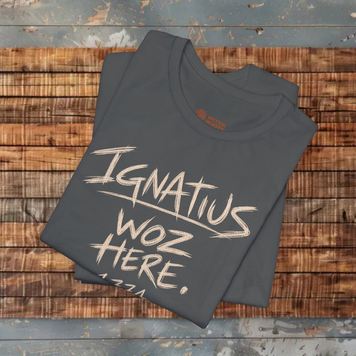 A folded dark gray t-shirt with a white hand-drawn style graphic reading 'Ignatius Woz Here, 1771' lies on a rustic wooden plank surface.