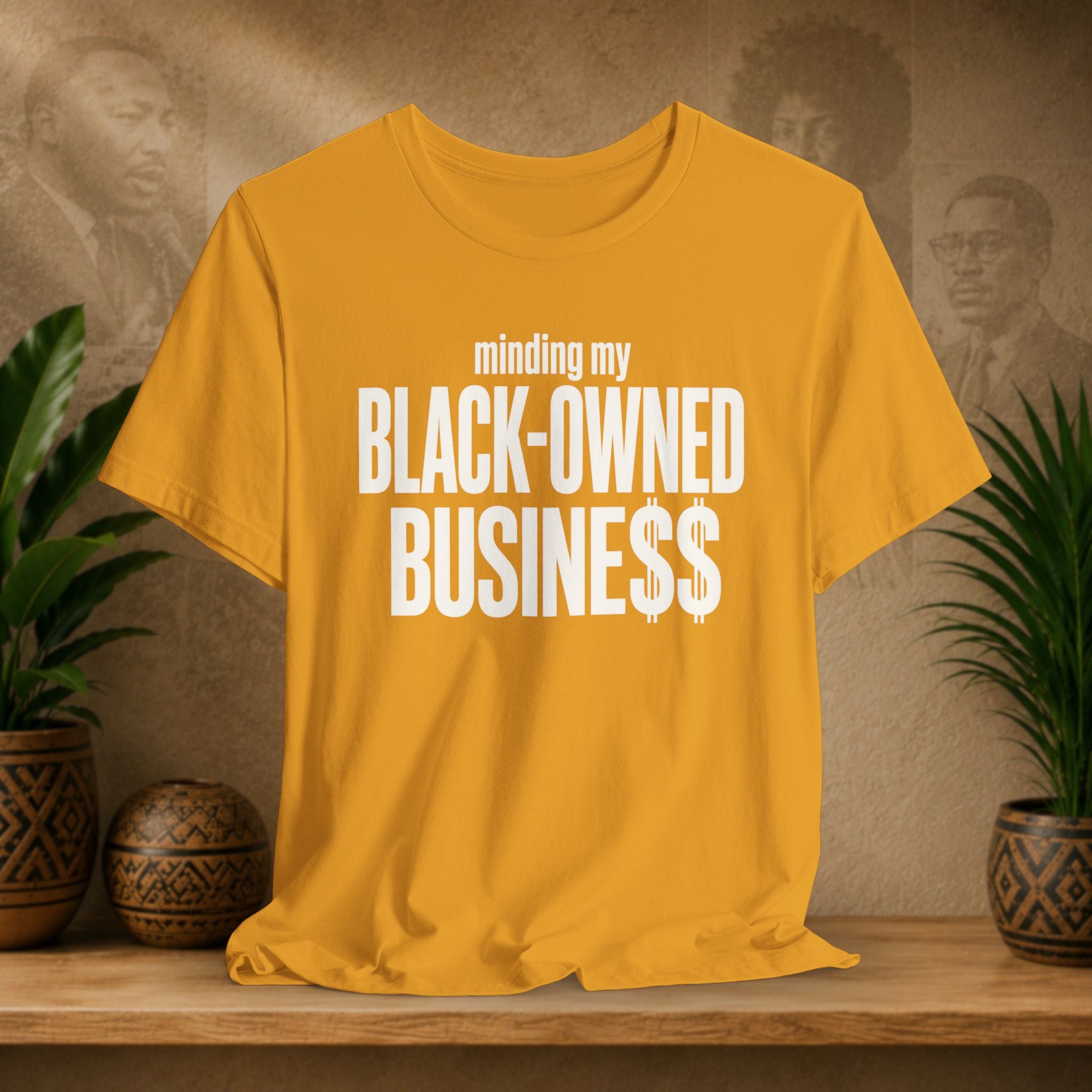 A mustard yellow short-sleeve t-shirt with white text reading 'minding my BLACK-OWNED BUSINESS$$' displayed on a wooden shelf with potted plants in the background.