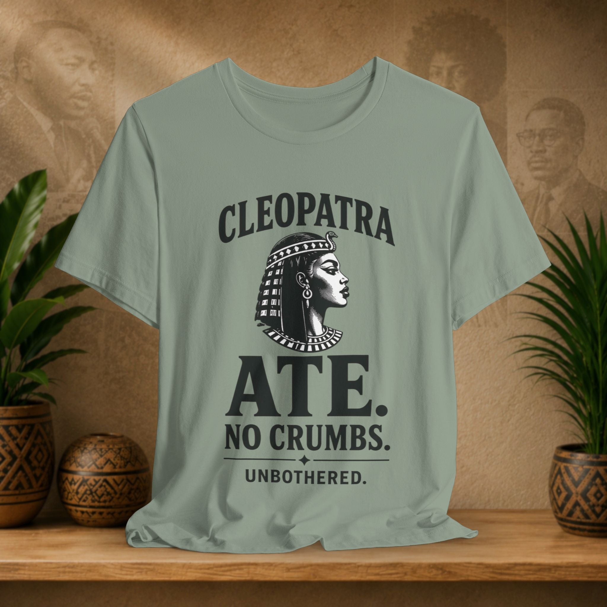 Light green t-shirt with Cleopatra graphic and text on a wooden surface with plants and a decorative item.