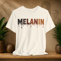 A beige short-sleeve crewneck t-shirt with the word 'MELANIN' printed across the chest in a dripping paint style, transitioning from black to brown. The shirt is displayed on a wooden shelf flanked by potted green plants against a textured brown background with sepia portraits.