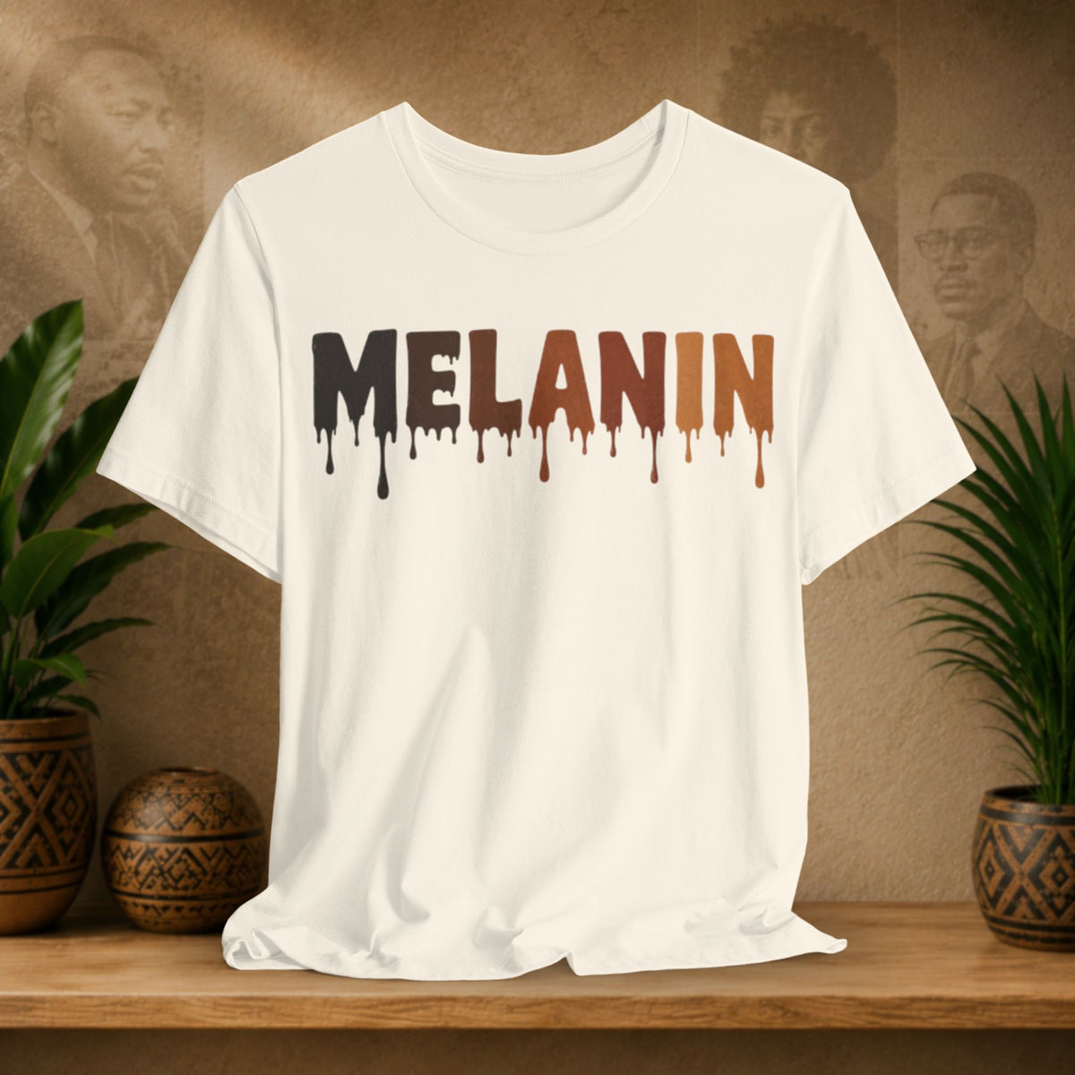 A beige short-sleeve crewneck t-shirt with the word 'MELANIN' printed across the chest in a dripping paint style, transitioning from black to brown. The shirt is displayed on a wooden shelf flanked by potted green plants against a textured brown background with sepia portraits.