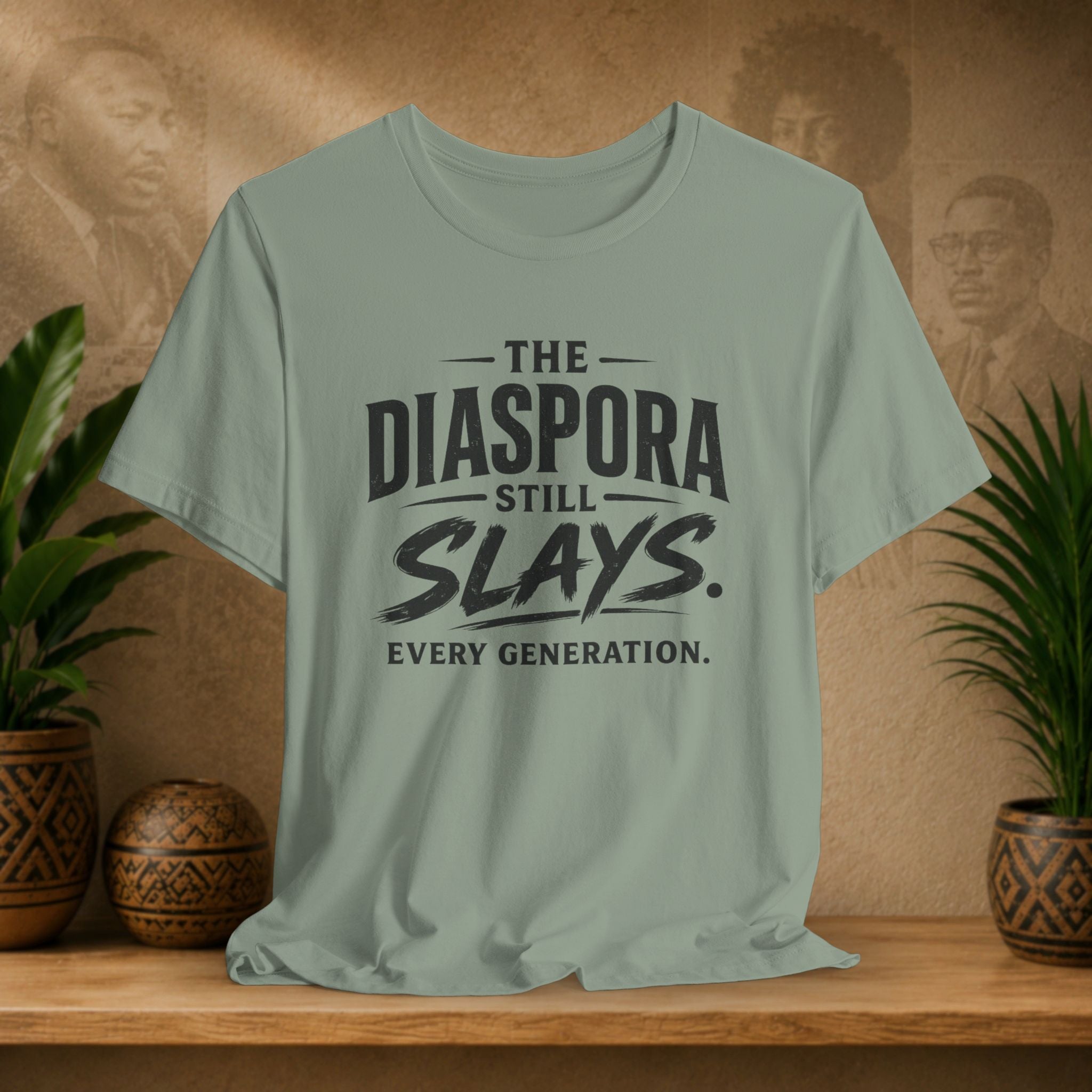 Green t-shirt with 'The Diaspora Still Slays Every Generation' text on a wooden surface with plants and a blurred background.