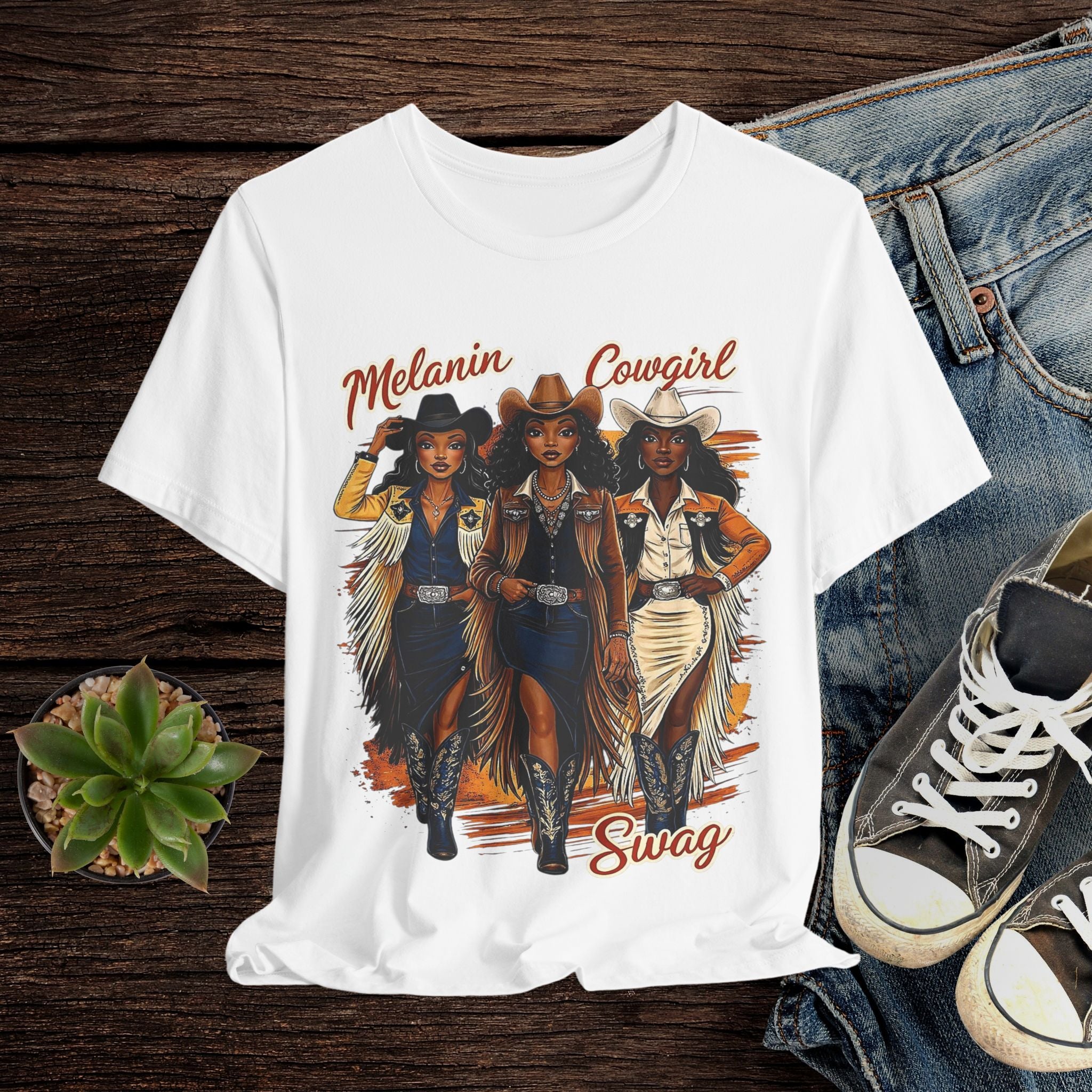 White t-shirt with 'Melanin Cowgirl Swag' design featuring three cowgirls on a wooden surface with jeans and sneakers.