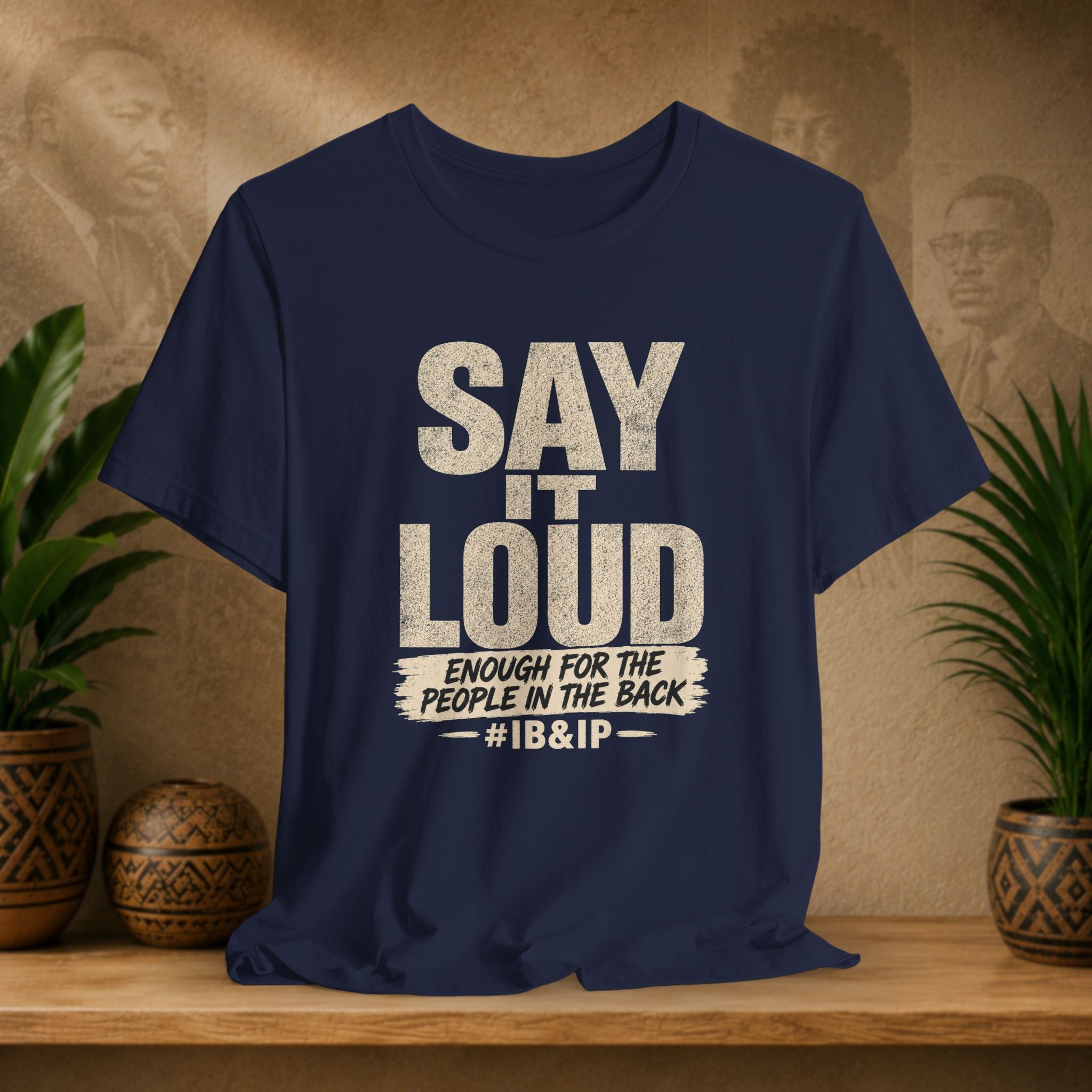 Navy t-shirt with text 'Say it Loud Enough for the People in the Back #IB&IP' on a wooden surface with plants and a textured wall background.