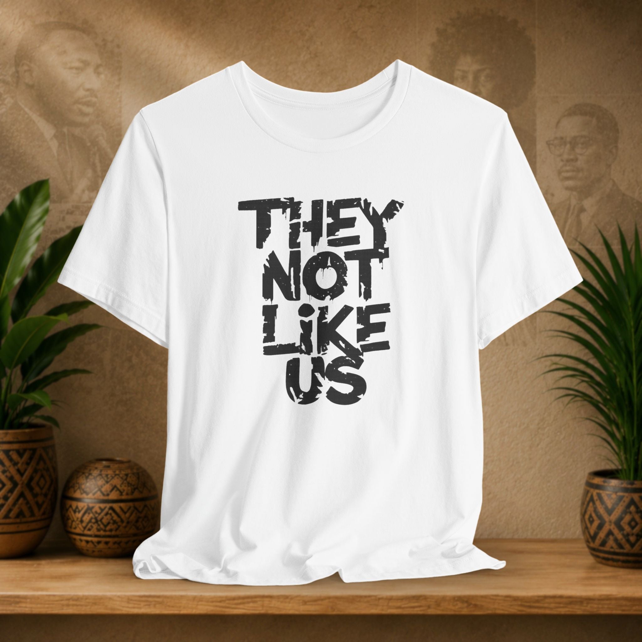 White t-shirt with 'THEY NOT LIKE US' text on a wooden surface with plants and a textured wall background