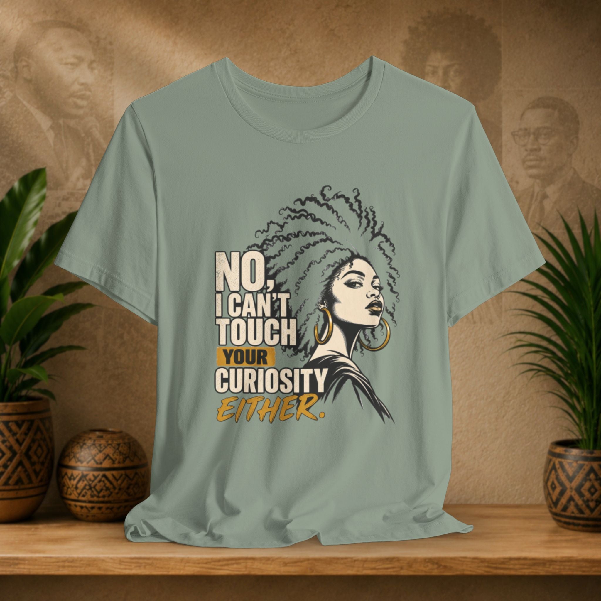 A sage green graphic t-shirt displayed on a wooden shelf, flanked by potted plants. The shirt features a stylized illustration of a woman with an afro and the text 'NO, I CAN'T TOUCH YOUR CURIOSITY EITHER.