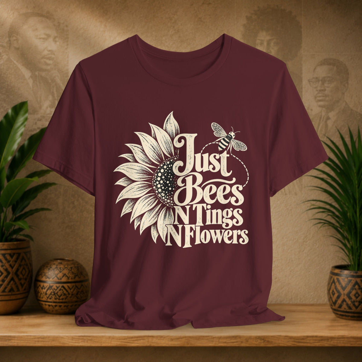 A maroon short-sleeve t-shirt with a cream-colored graphic that reads 'Just Bees N Tings N Flowers' alongside a sunflower and bee illustration, displayed on a wooden shelf with plants in the background.