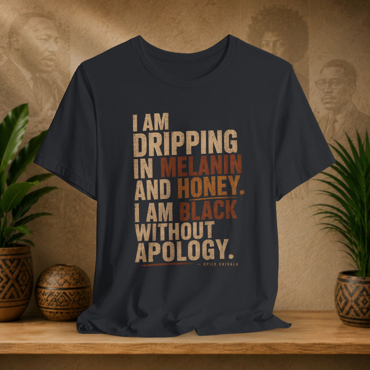 A black graphic t-shirt displayed on a wooden shelf, flanked by potted plants. The shirt features a typographic design in beige and brown text referencing Aipil Enochala's quote about dripping melanin and honey.