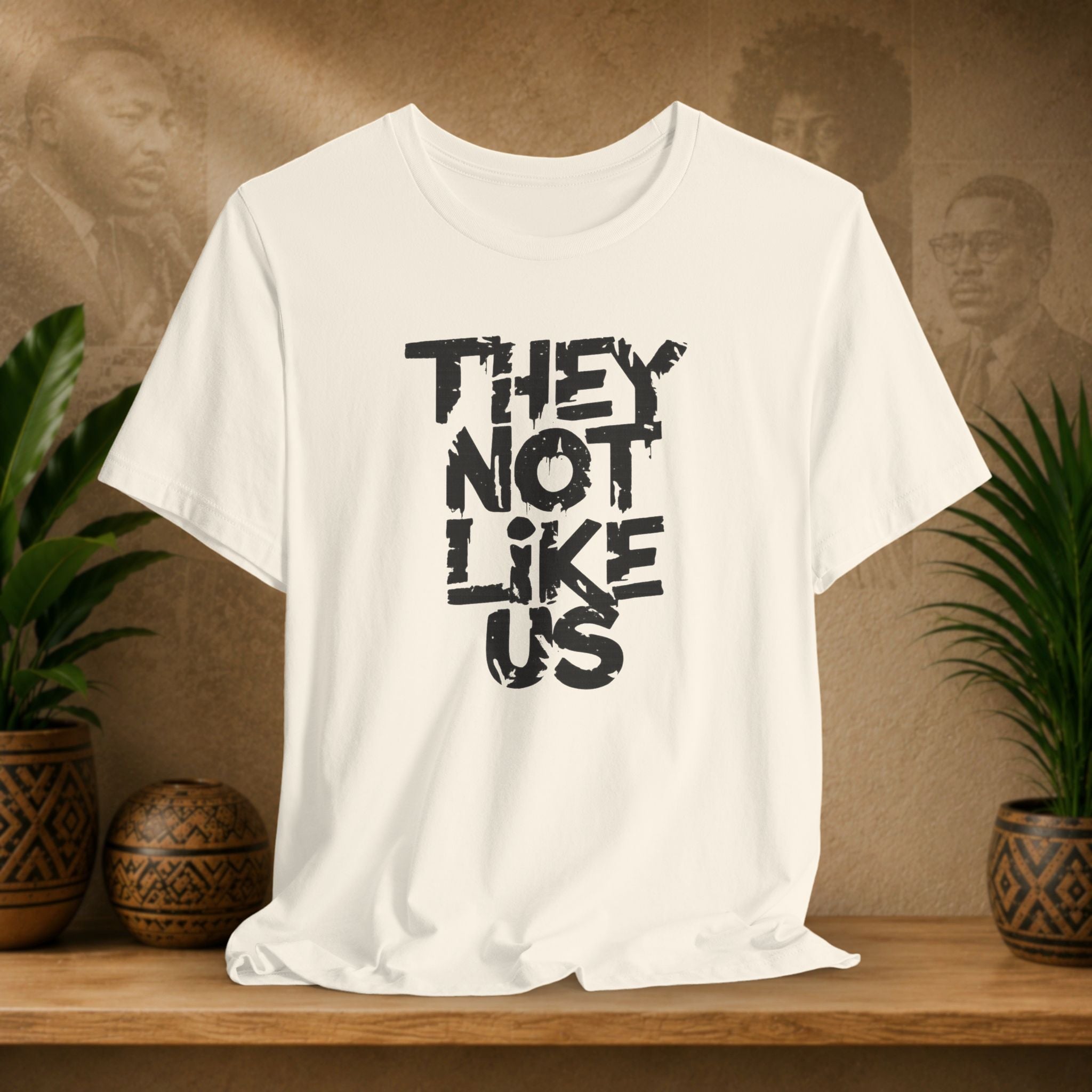 Beige t-shirt with 'THEY NOT LIKE US' text on a wooden surface with plants and a textured wall background
