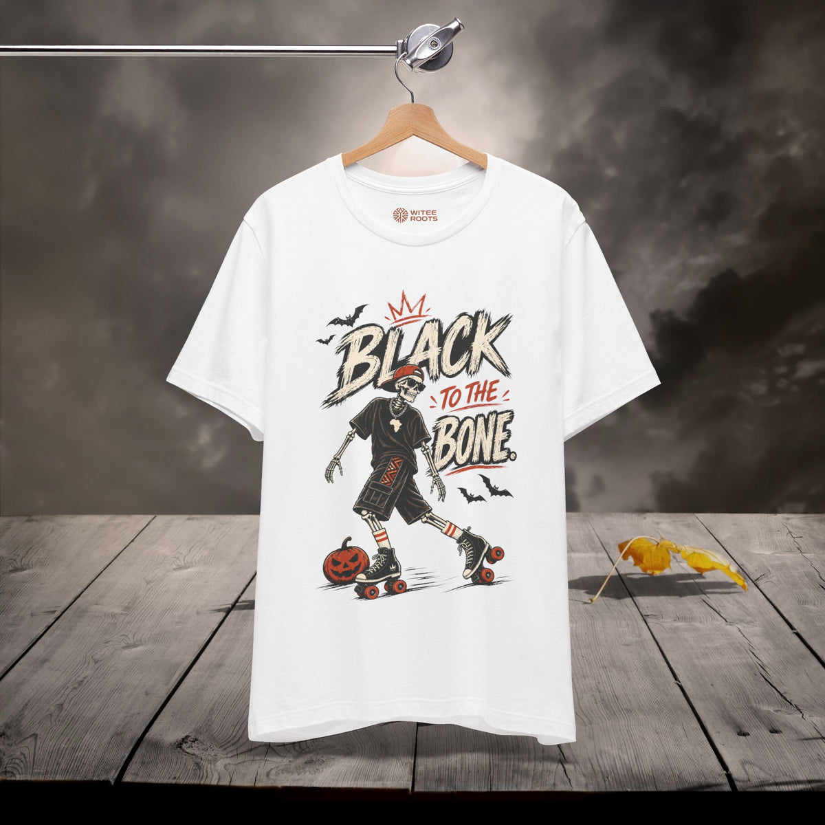 A white graphic t-shirt hanging on a wooden hanger against a dark, smoky background with a wooden plank floor. The shirt features a Halloween-themed graphic with the text 'BLACK TO THE BONE' and an illustration of a skeleton roller skate.