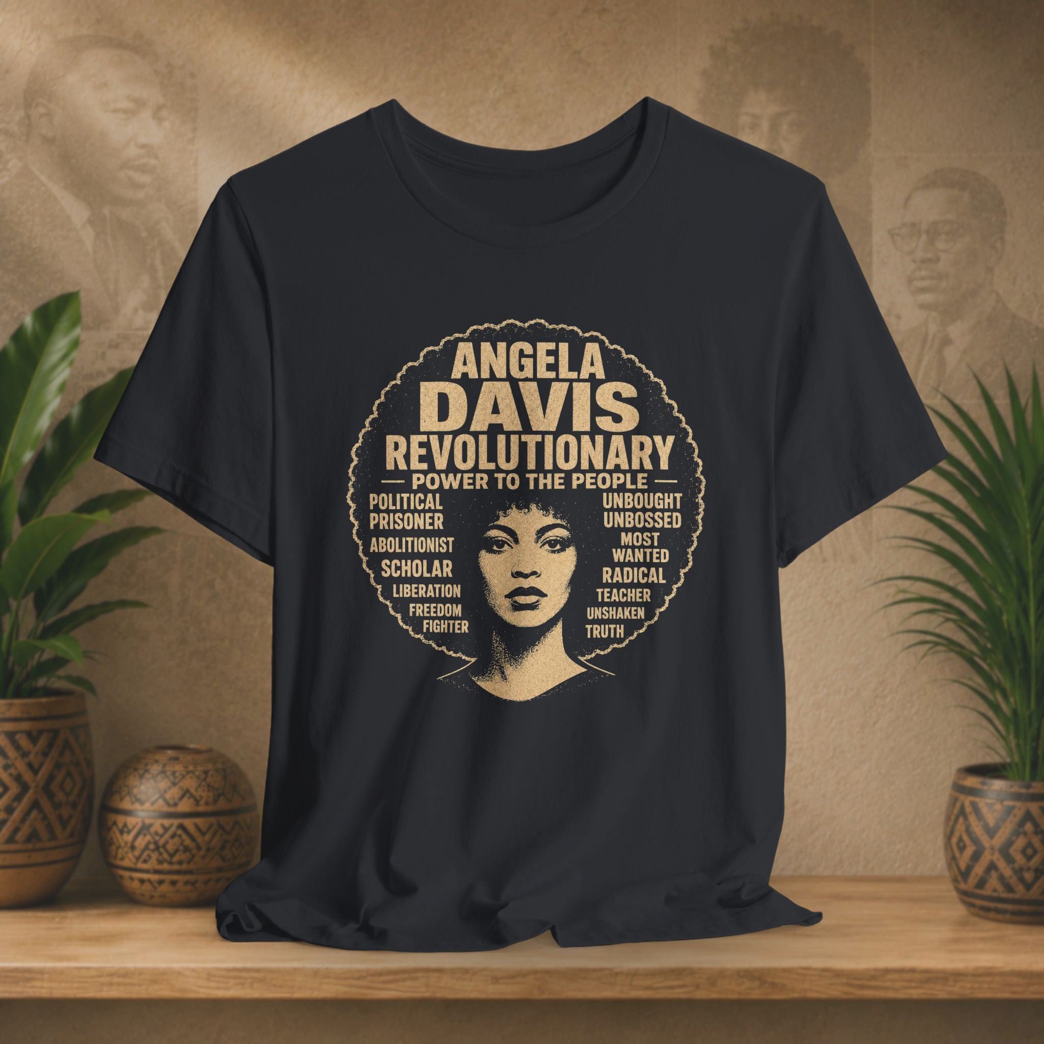 A black short-sleeve graphic T-shirt displayed on a wooden surface with potted plants and a warm textured backdrop. The shirt features a large circular design with the text 'ANGELA DAVIS REVOLUTIONARY' and a stylized portrait of Angela Davis in the center.