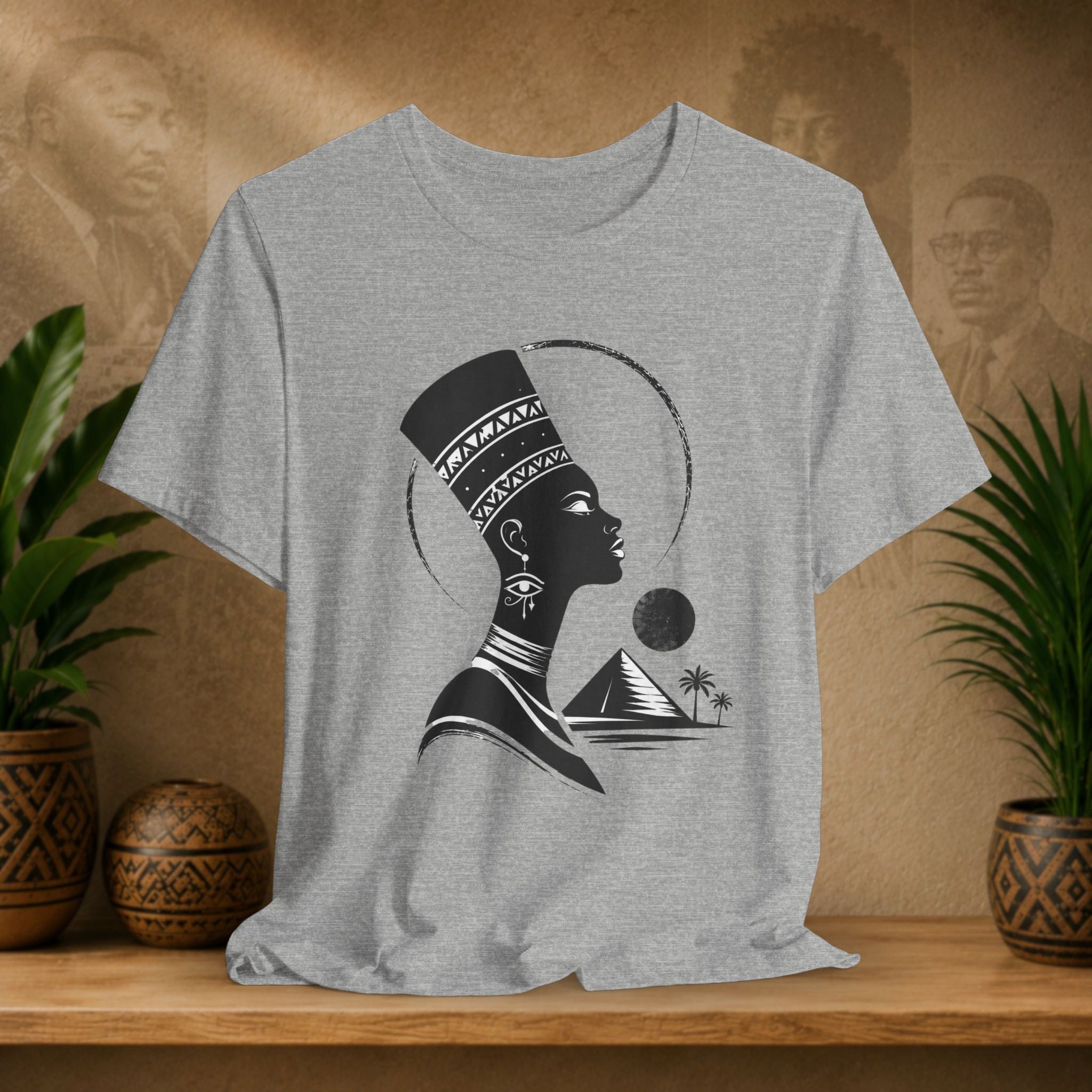 Gray t-shirt with a black silhouette design of a woman with a headdress and pyramid on a wooden shelf with plants and decorative items.