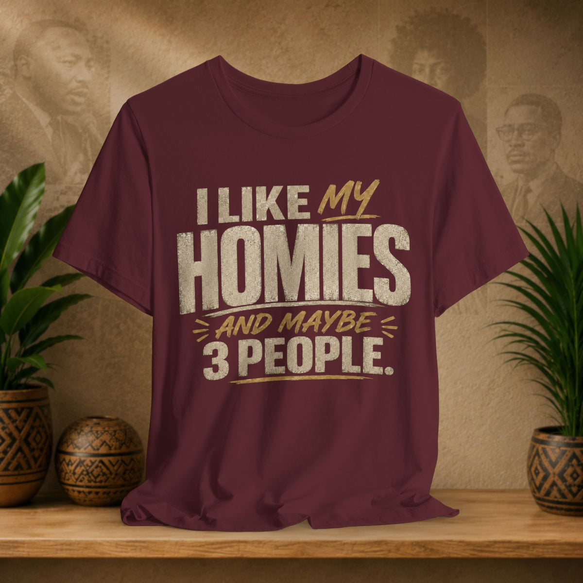 A maroon graphic t-shirt displayed on a wooden shelf. The shirt features a humorous slogan printed in white and tan lettering: 'I LIKE MY HOMIES and maybe 3 PEOPLE.