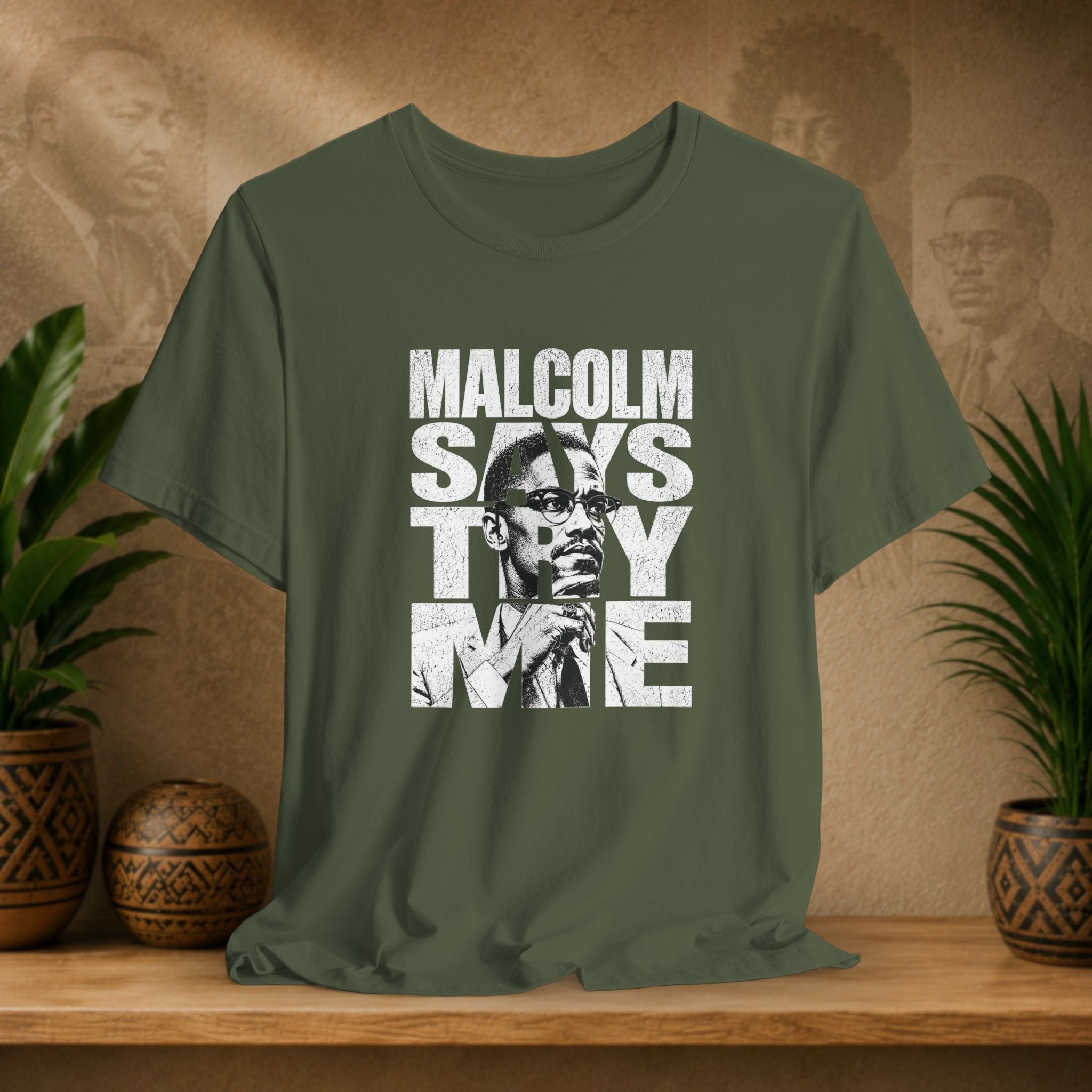 Green t-shirt with 'Malcolm Says Thy Me' graphic on a wooden surface with plants and a textured wall in the background.