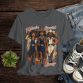 Gray t-shirt with 'Melanin Cowgirl Swag' design featuring three cowgirls, displayed with jeans and sneakers on a wooden surface.