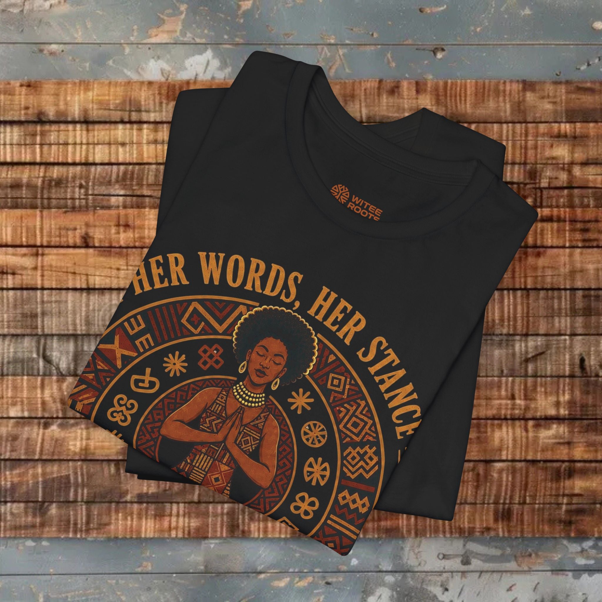 A folded brown t-shirt with a graphic print of a Black woman and the text 'HER WORDS HER STANCE' in gold, displayed on a rustic wooden plank background.