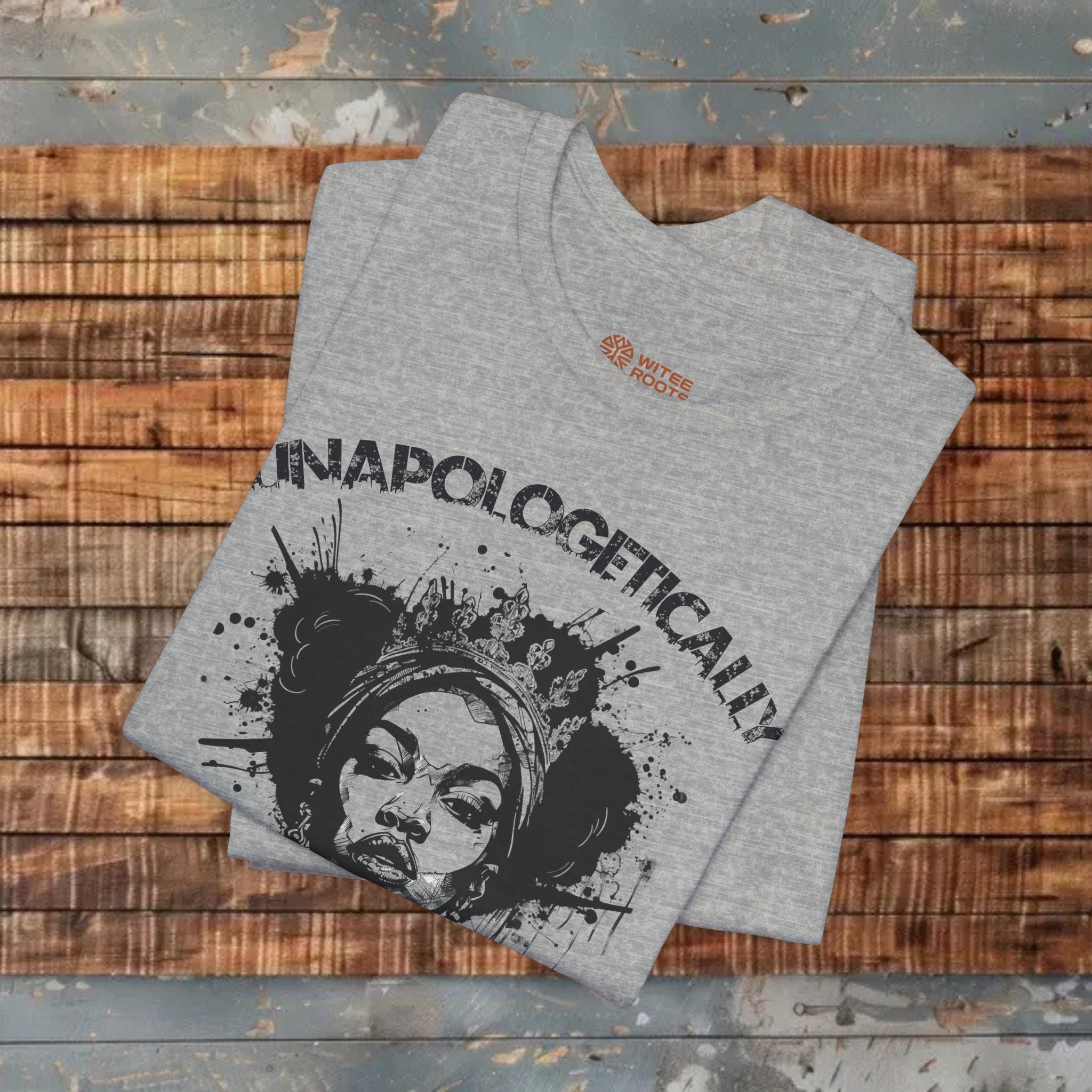 A folded heather gray t-shirt with a black graphic print reading 'UNAPOLOGETICALLY' and a stylized face illustration, displayed on a rustic wooden plank background.