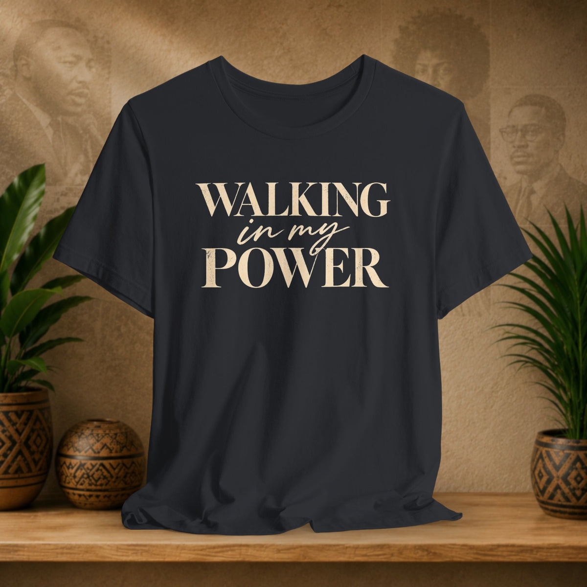 A black t-shirt with the text 'WALKING in my POWER' printed in beige on the front, displayed on a wooden shelf with decorative plants and a textured background.
