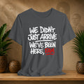 Gray t-shirt with text 'WE DIDN'T JUST ARRIVE WE'VE BEEN HERE, FAM' on a wooden surface with plants and a textured wall background.