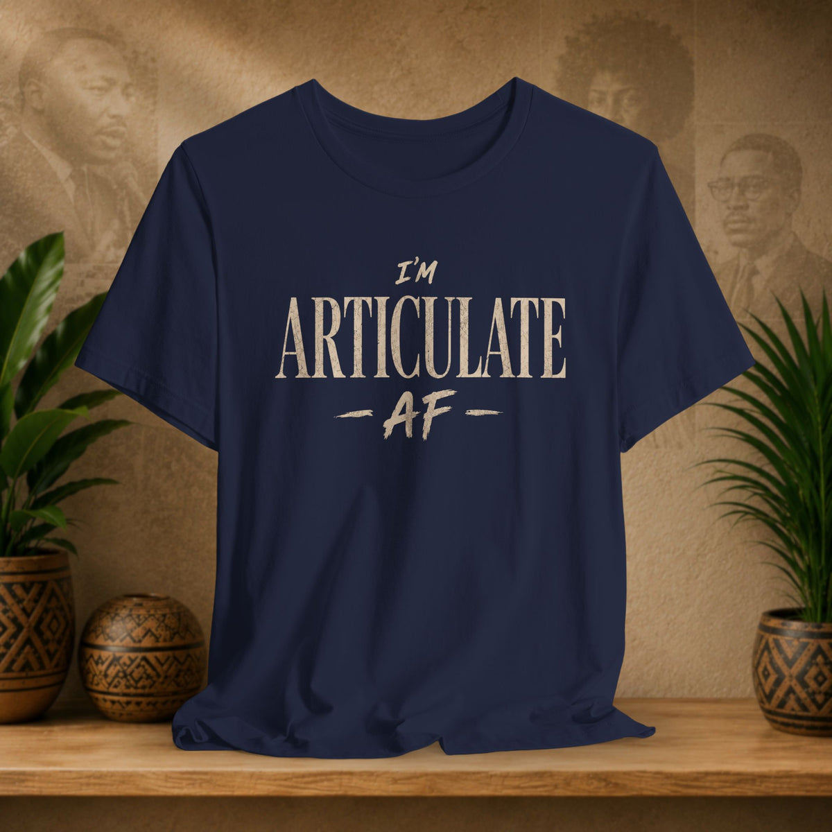 A navy blue short-sleeve t-shirt with the text 'I'M ARTICULATE AF' printed in a distressed cream font, displayed on a wooden shelf with potted plants and a textured background.