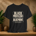 Black t-shirt with 'Black Privilege: Being the Blueprint' text on a wooden surface with plants and a textured wall background.