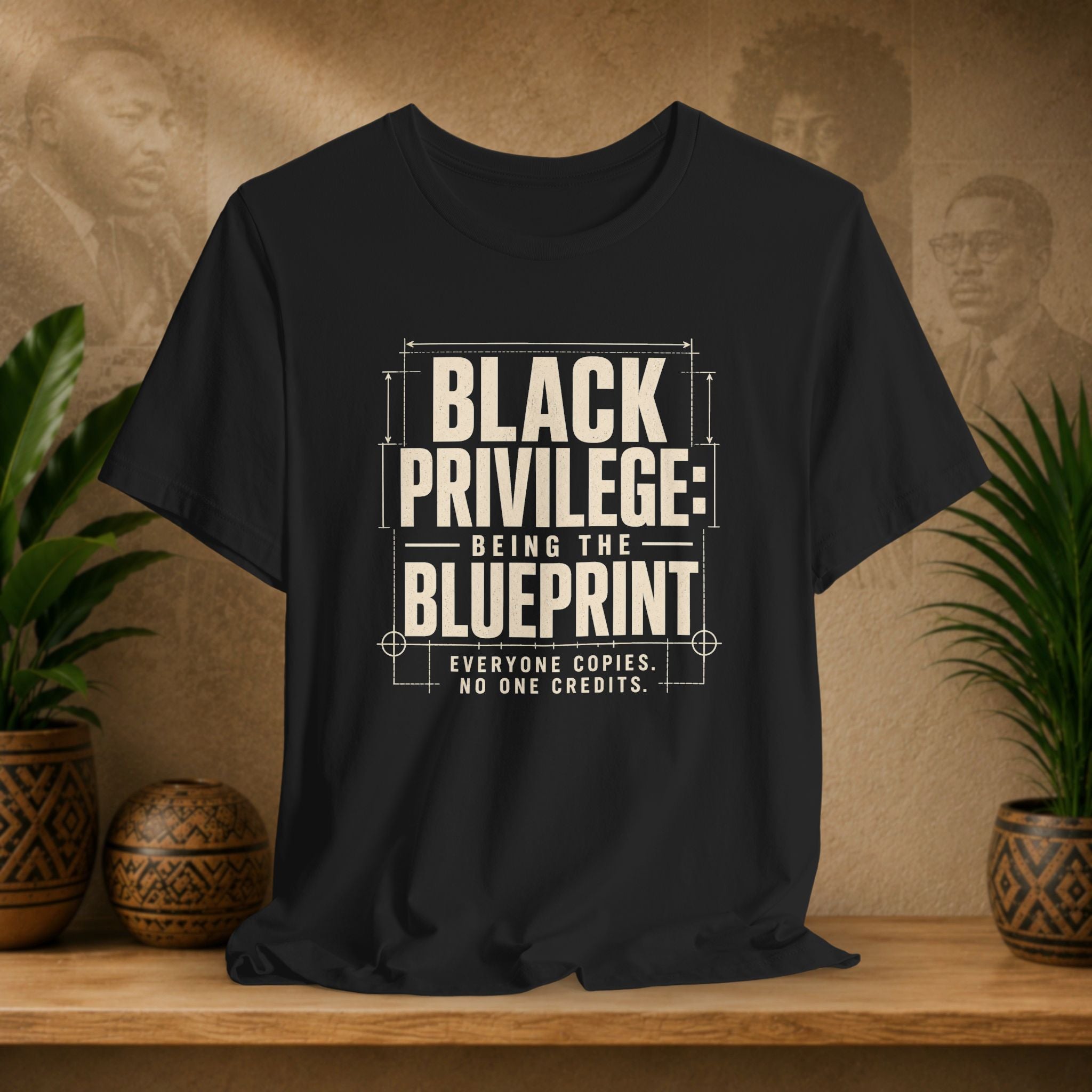 Black t-shirt with 'Black Privilege: Being the Blueprint' text on a wooden surface with plants and a textured wall background.