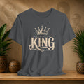 Gray t-shirt with 'Black is King' design on a wooden surface with plants and decorative items in the background.