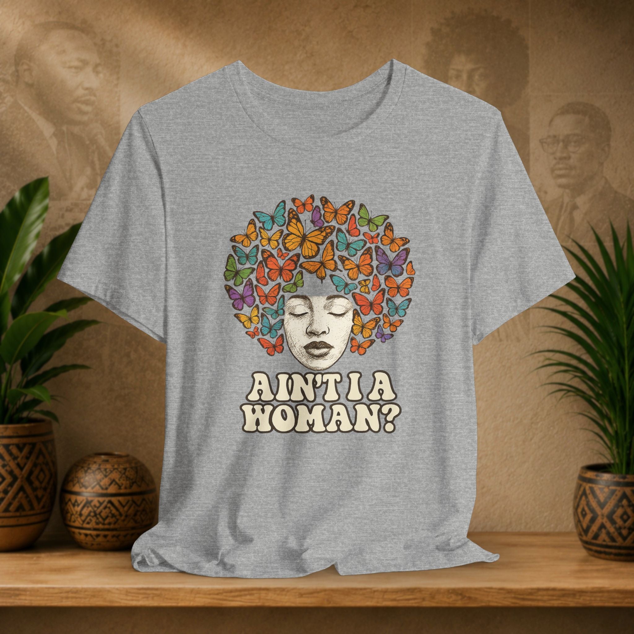 Gray t-shirt with a colorful butterfly design and text on a wooden shelf with plants and decorative items.