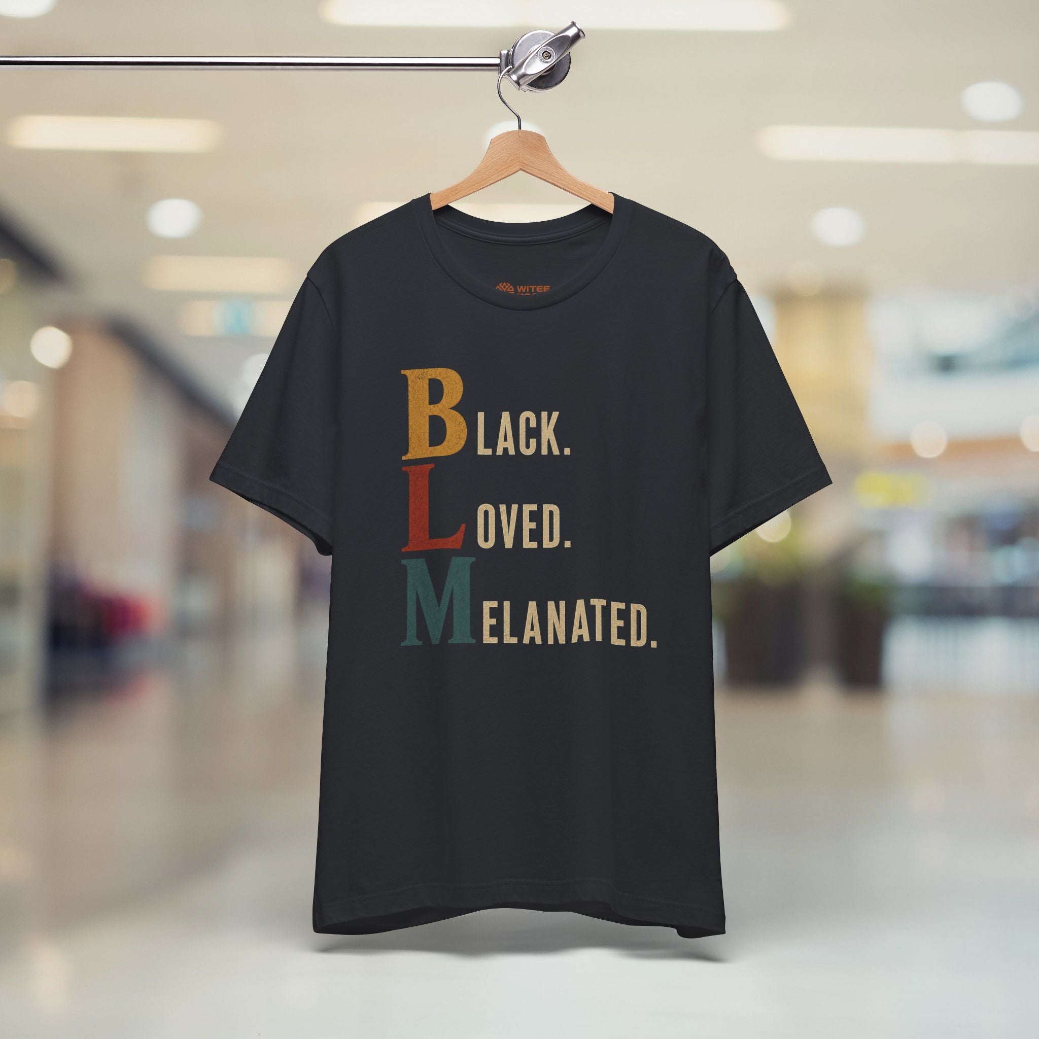 A black short-sleeve graphic T-shirt hanging on a wooden hanger against a blurred indoor shopping mall background. The shirt features the text 'BLACK.