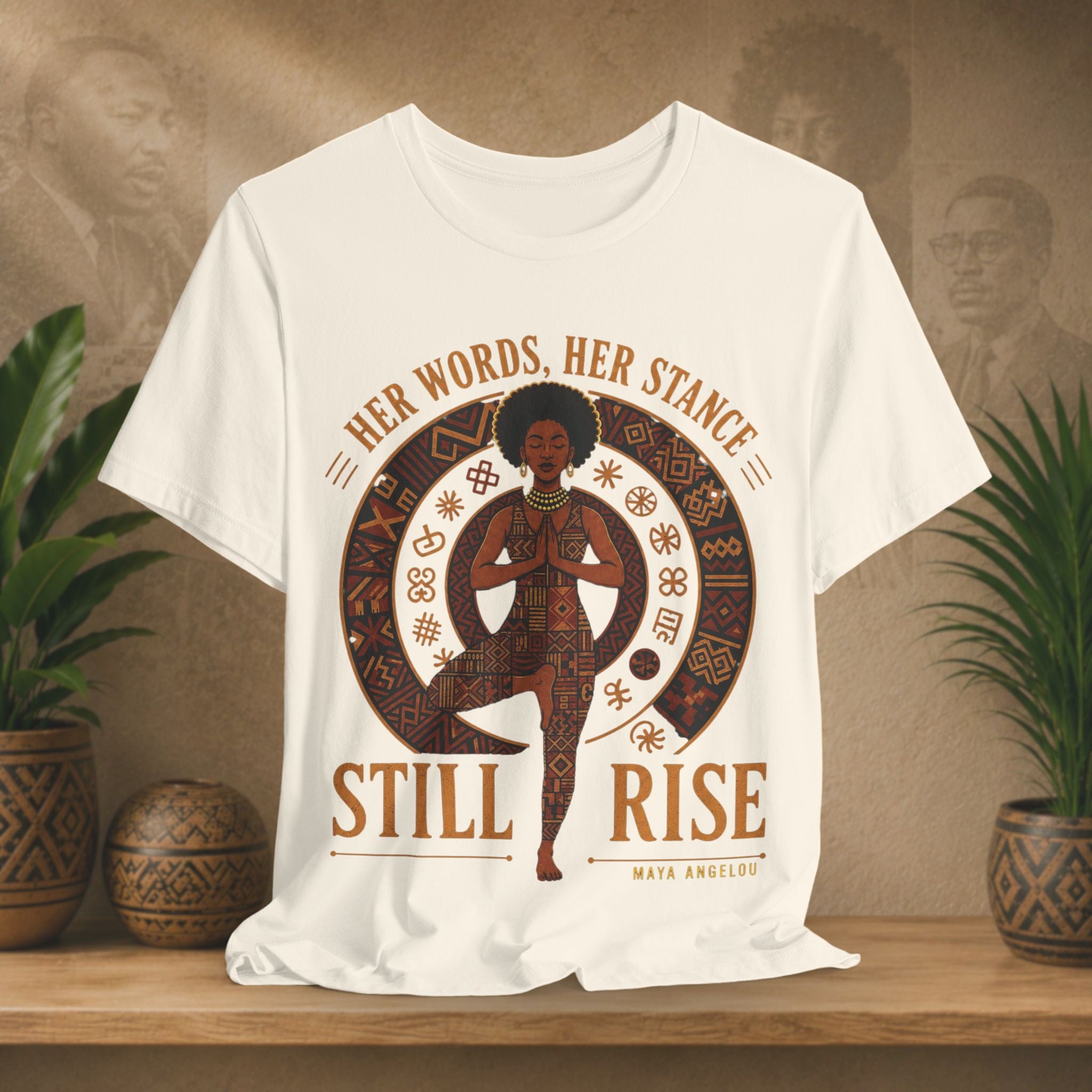 A beige short-sleeve graphic T-shirt displayed on a wooden surface with potted plants and a warm textured backdrop. The shirt features a large central circular design with an illustrated figure in a yoga pose, surrounded by brown decorative motifs and the text 'HER WORDS, HER STANCE STILL RISE' with 'MAYA ANGELUS' below.