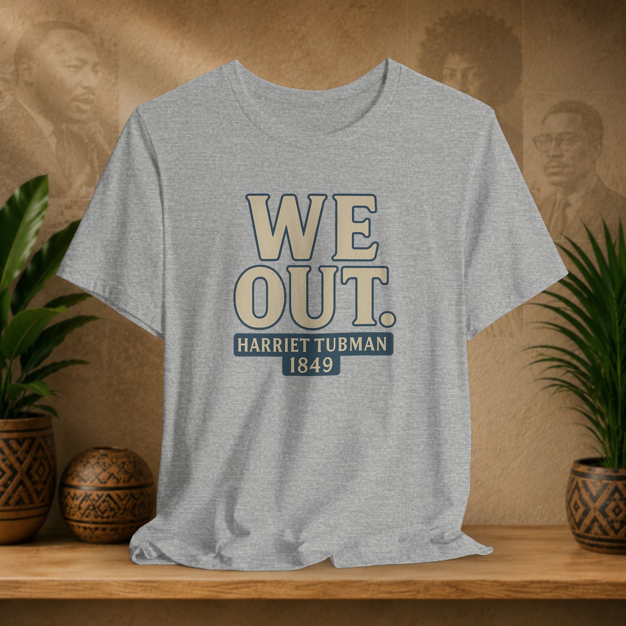 Gray t-shirt with 'WE OUT. HARRETT TUBMAN 1849' text on a wooden shelf with plants and a blurred background.