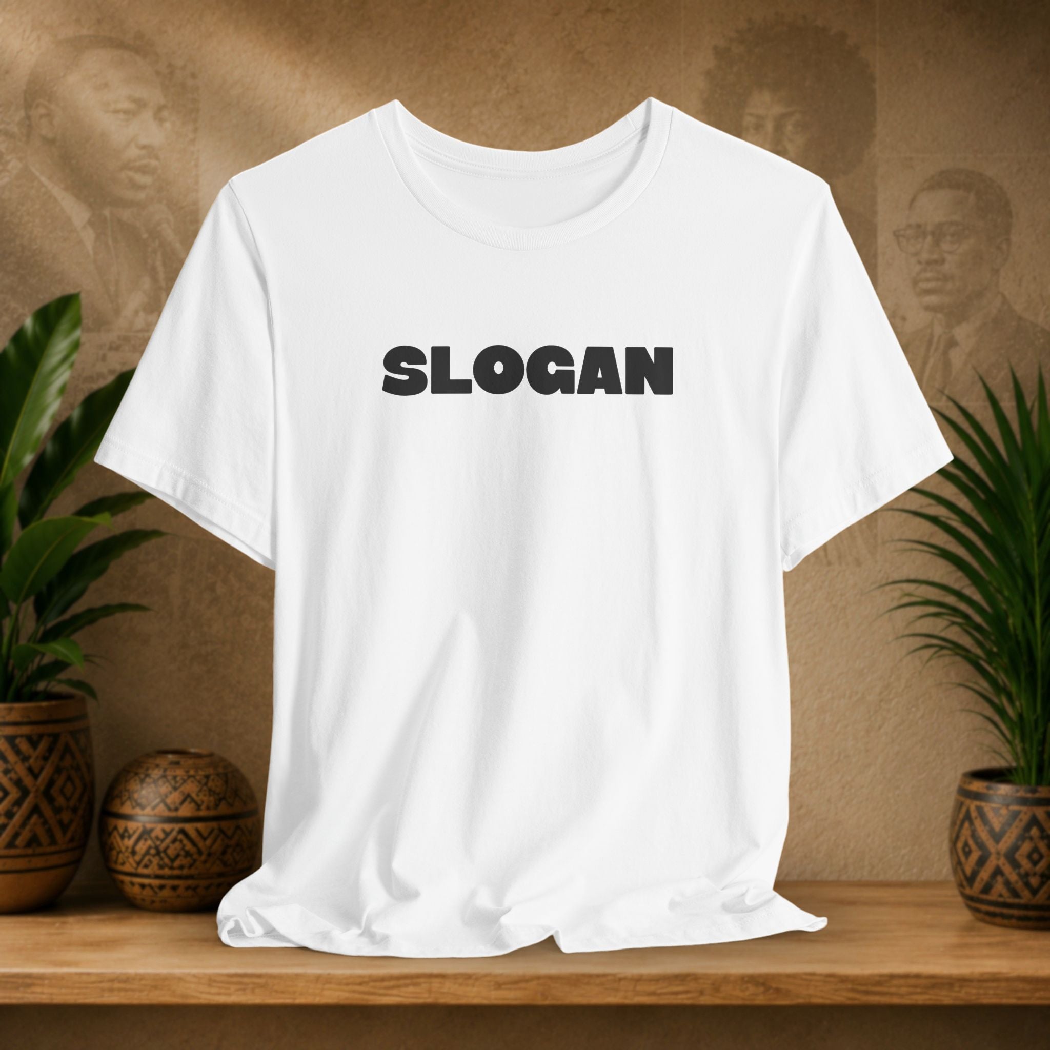 A white short-sleeve crewneck t-shirt with 'YOUR SLOGAN HERE' printed in distressed black text on the chest, displayed on a wooden shelf with potted plants in the background.