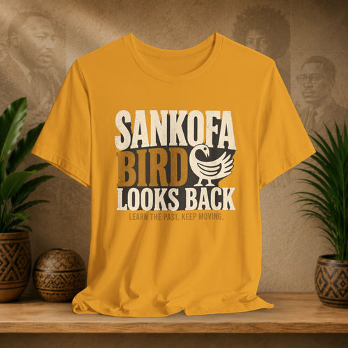 Yellow t-shirt with 'Sankofa Bird Looks Back' design on a wooden surface with plants and a textured wall background.