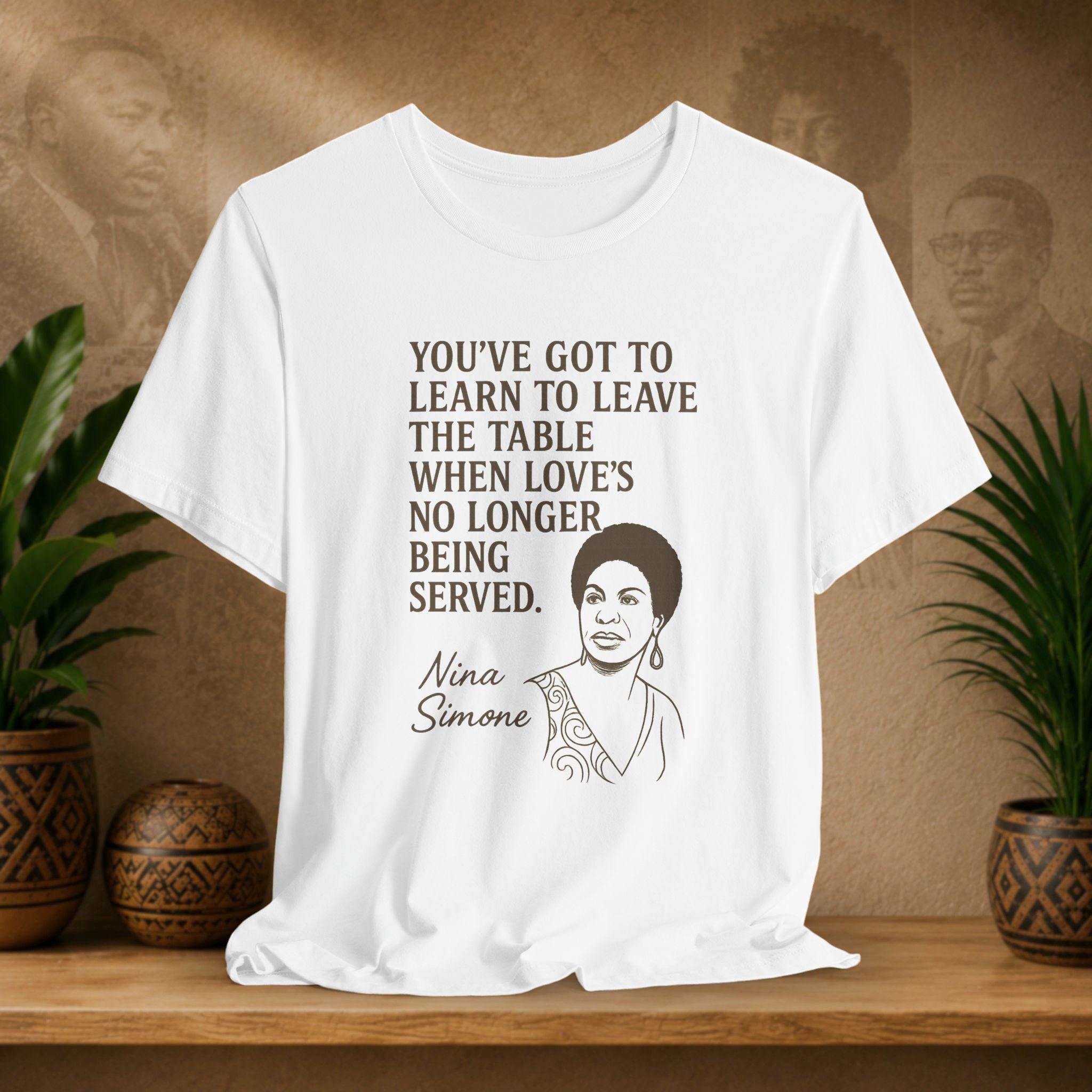 White t-shirt with Nina Simone quote on a wooden shelf with plants and a brown background