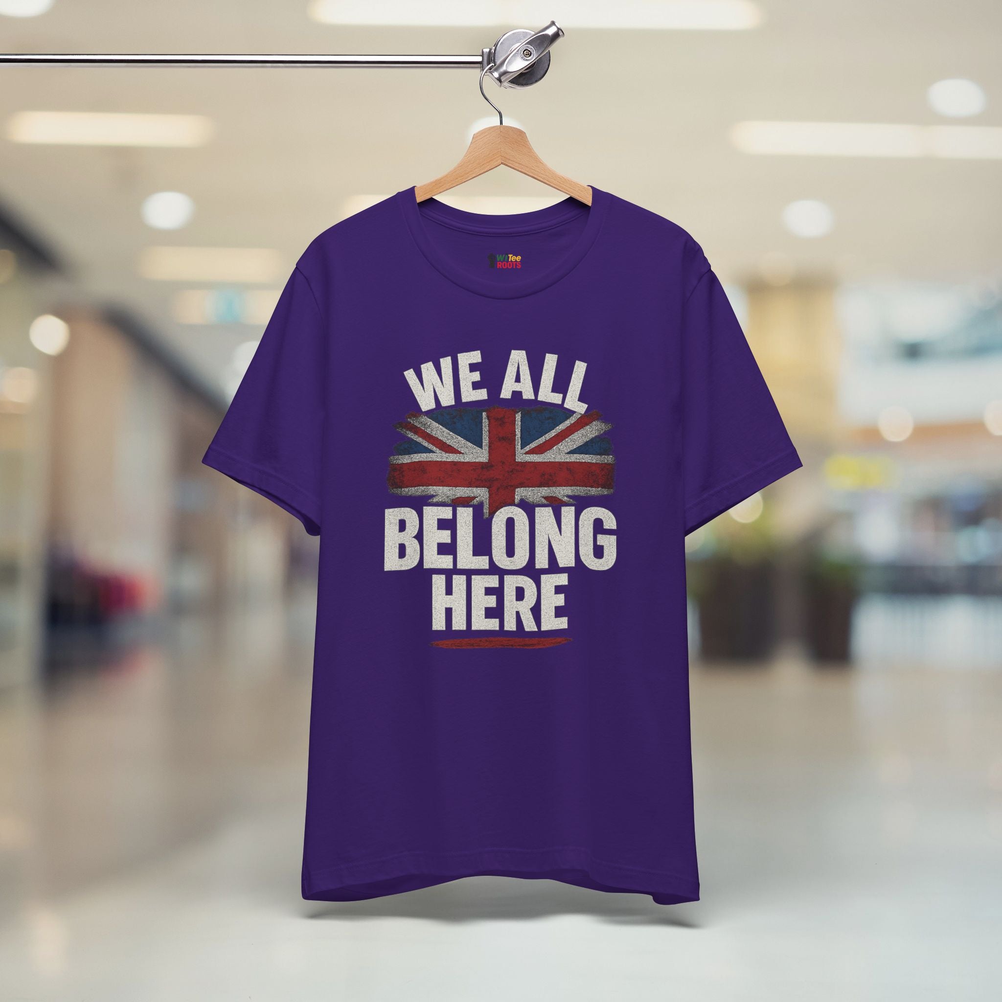 Purple t-shirt with a Union Jack design and text 'WE ALL BELONG HERE' on a hanger in a store.