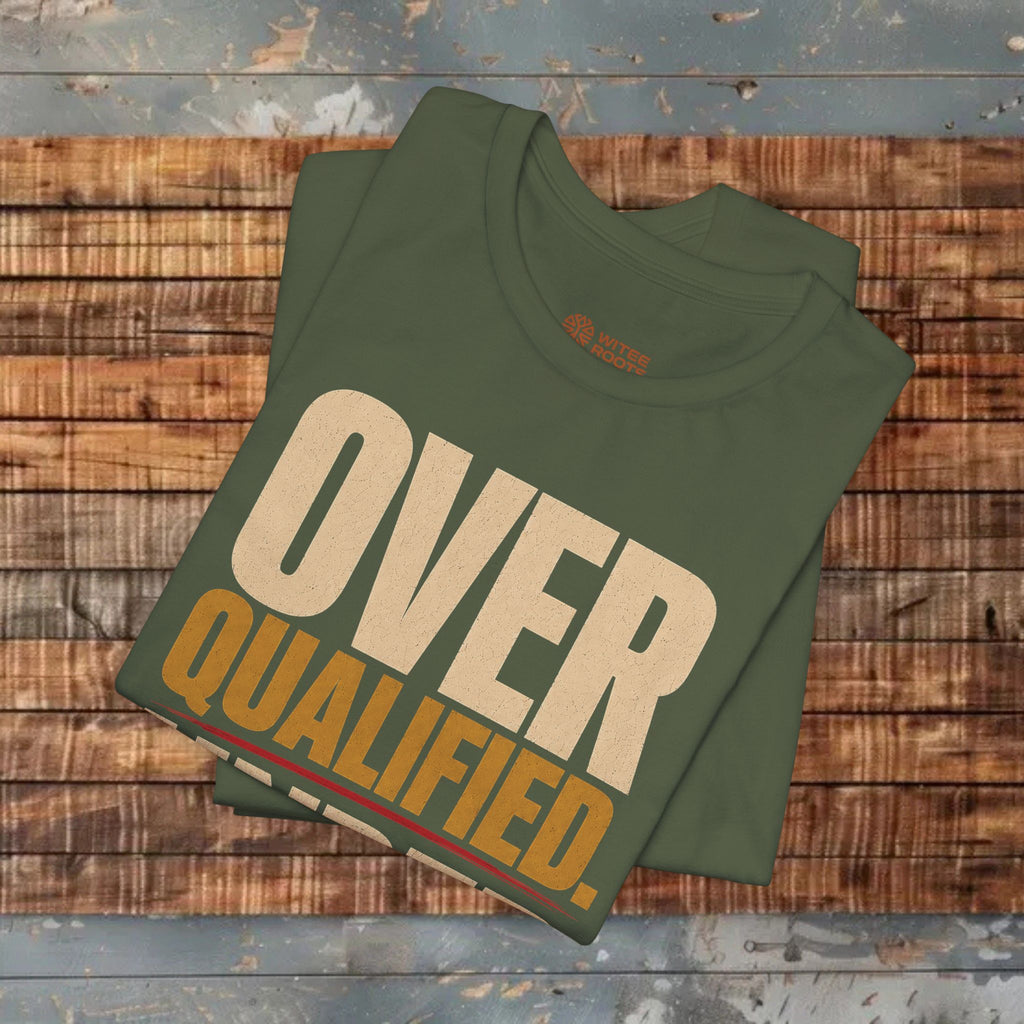 A folded olive green t-shirt with a retro-style graphic reading 'OVER QUALIFIED.' in beige and mustard yellow, displayed on a rustic wooden plank background.