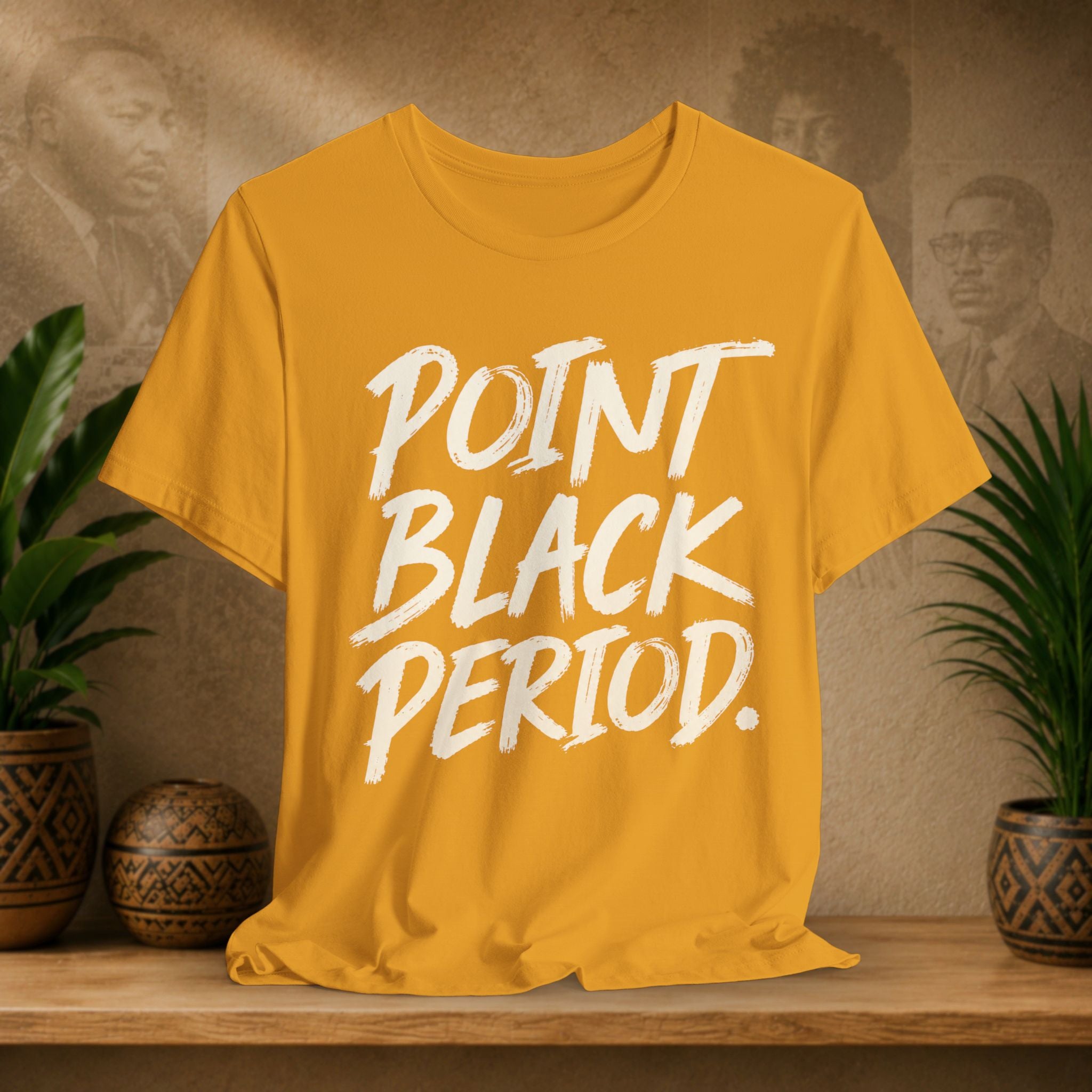 A mustard yellow t-shirt with the text 'POINT BLACK PERIOD.' printed in a white brush-stroke font, displayed on a wooden shelf with potted plants and a textured background.